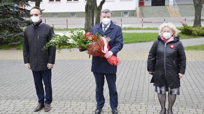 zdjęcie przedstawia trzy osoby: w środku burmistrz niosący wiązankę biało-czerwonych kwiatów, po prawej wiceburmistrz, po prawej wicestarosta
