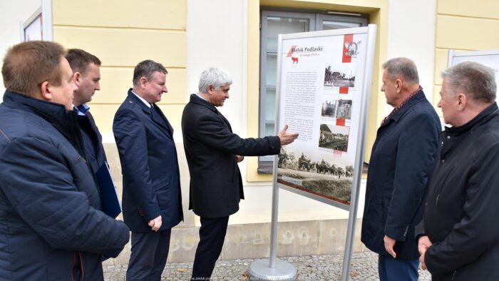 Ilustracja przedstawia sześciu mężczyzn stojących wokół tablicy wystawowej poświęconej miastu Bielsk Podlaski.