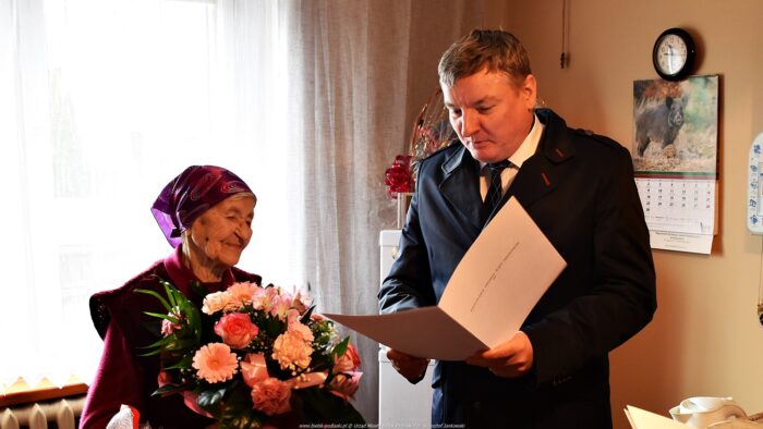 Zdjęcie przedstawia dwie osoby: stulatkę i burmistrza. Stulatka trzyma bukiet kwiatów, burmistrz odczytuje list gratulacyjny. W tle wnętrze drewnianego domu jubilatki.