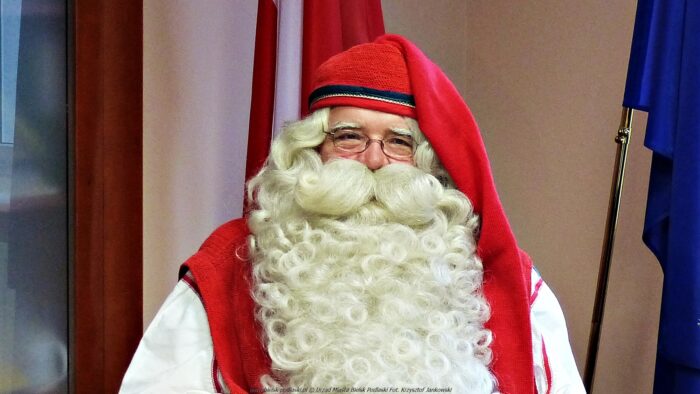 Zdjęcie przedstawia prawdziwego Mikołaja z Laponii podczas wizyty w Urzędzie Miasta Bielsk Podlaski w 2019 roku. Mikołaj siedzi na tle flag: Polski i Unii Europejskiej.