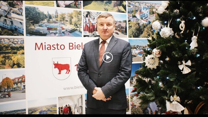 Ilustracja przedstawia slajd z filmu z życzeniami: burmistrza stojącego na tle ścianki promocyjnej miasta Bielsk Podlaski. Po prawej stronie kadru widać fragment świątecznej choinki.