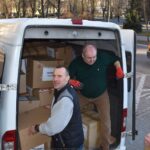 Ilustracja przedstawia pracowników urzędu miasta podczas ładowania darów do furgonetki.
