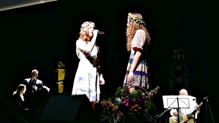 Dwie śpiewające kobiety z wiankami we włosach na scenie BDK-u. Stoją bokiem do widowni, zwrócone do siebie twarzami. W tle muzycy z instrumentami, przed nimi kosz z kwiatami.