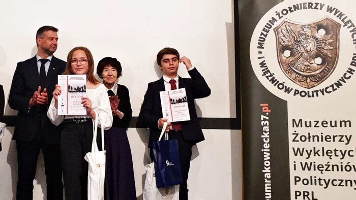 Zdjęcie przedstawia 4 osoby: dwie dorosłe i dwoje młodych laureatów. Laureaci trzymają przed sobą dyplomy, a starsza kobieta - wdowa po rotmistrzu - bije brawo. Z prawej stoi rollup z logo Muzeum Żołnierzy Wyklętych i Więźniów Politycznych.