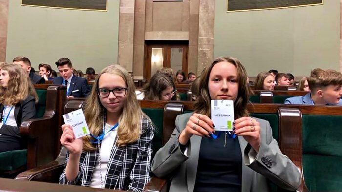 Dwie nastoletnie posłanki Kornelia i Aleksandra siedzą w ławach sejmowych wśród innych młodych ludzi z całej Polski. Przed sobą trzymają plakietki - identyfikatory.