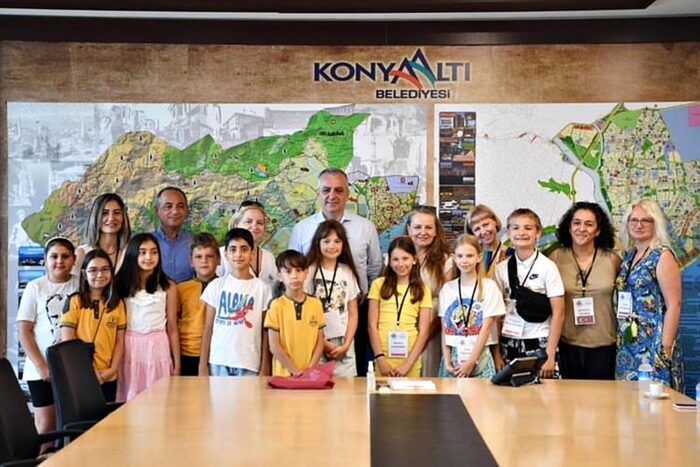 Zdjęcie przedstawia 18 osób stojących na tle dwóch map na ścianie i napisu "konyaalti". Na pierwszym planie stoją dzieci, w tle dorośli - opiekunowie. Zdjęcie zrobiono w sali konferencyjnej.