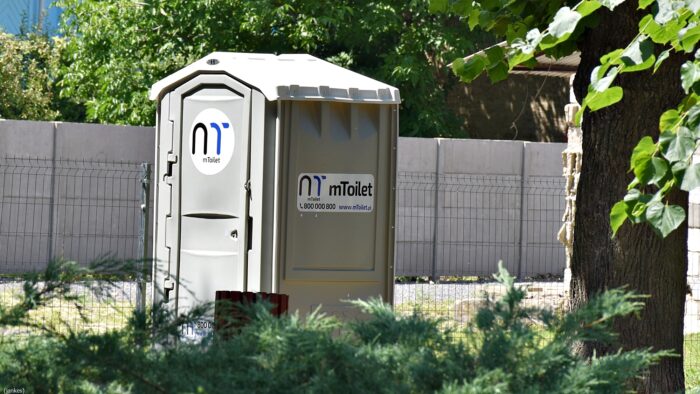 Nowa toaleta przenośna toi na tle płotu i w otoczeniu zieleni bielskiego parku.