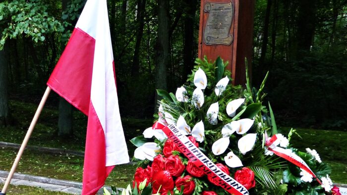 Zdjęcie przedstawia fragment krzyża ustawionego na miejscu zbrodni w Lesie Pilickim. Przed krzyżem leży wieniec w kolorze biało-czerwonym oraz wisi flaga narodowa.