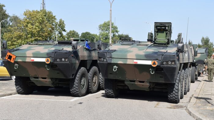Dwa wojskowe pojazdy opancerzone stoją na parkingu przed dworcem. Za nimi, w dwóch rzędach, ustawione są kolejne pojazdy wojskowe.