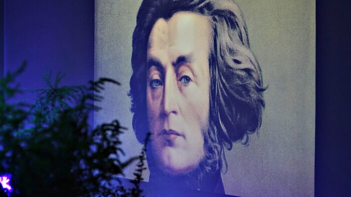 Obraz mężczyzny - Adama Mickiewicza - wyświetlony na ścianie z lewej strony kępa roślin.