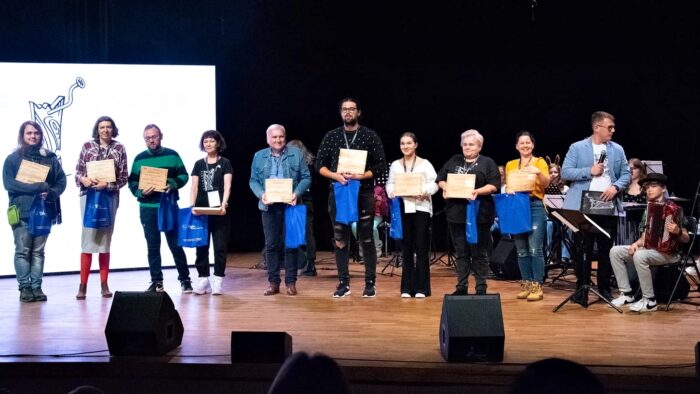 Dziesięć osób - laureatów festiwalu - ustawionych w rzędzie na scenie domu kultury. W rękach trzymają nagrody - statuetki, w kształcie drewnianych desek i niebieskie reklamówki z gadżetami promocyjnymi miasta Bielsk Podlaski.
