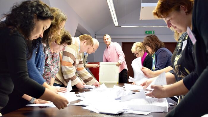 Ilustracja przedstawia grupę urzędników przeliczającą głosy. Kartki do głosowania są wysypane z urny na duży stół, wokół którego stoją przeliczający je urzędnicy.