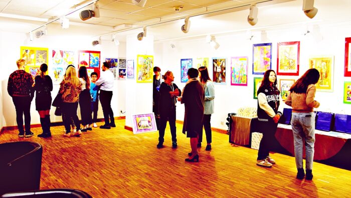 Grupa osób zwiedzających wystawę prac konkursowych podczas biennale. Kolorowe prace wywieszone są na ścianach galerii.