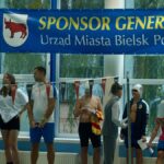 Grupa zawodników stojących na tle okna pływalni. Nad nimi baner z napisem "Sponsor Generalny Urząd Miasta Bielsk Podlaski".