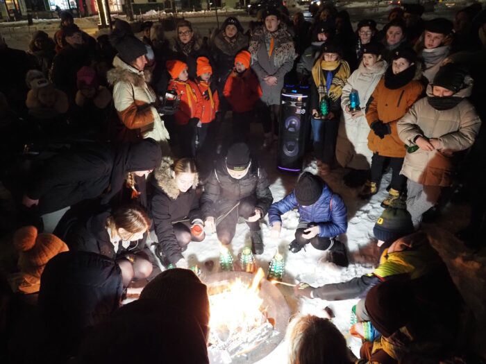 Zdjęcie przedstawia uczestników uroczystości przekazania ognia z Betlejem społeczności harcerskiej oraz mieszkańcom Bielska Podlaskiego. Wokół ogniska znajdują się dzieci, które zapalają lampiony. Wokół nich stoją dorośli.