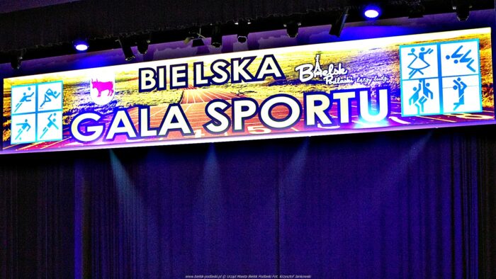 Ilustracja wyświetlona nad sceną domu kultury z napisem: "Bielska Gala Sportu", a także logotypem "Bielsk Podlaski łączy ludzi" i piktogramami symbolizującymi różne dyscypliny sportowe. W tle fioletowa kurtyna, na górze - lampy sceniczne.