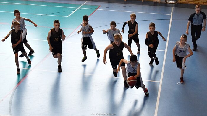 Grupa dzieci w strojach sportowych podczas gry w koszykówkę. Jeden z zawodników kozłuje piłkę, pozostali biegną za nim. Po prawej sędzia z gwizdkiem w ustach.