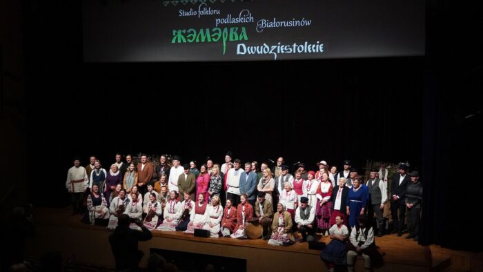 Kilkudziesięcioosobowa grupa osób stoi i kuca na scenie. Nad nimi wisi baner z napisem "Studio folkloru podlaskich Białorusinów".