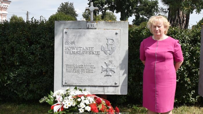 Zastępca burmistrza miasta stoi przy tablicy pamiętkowej, poświęconej Powstaniu Warszawskiemu. Pod tablicą leży wieniec kwiatów w barwach bieli i czerwieni. W tle zieleń cmentarna.