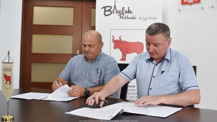 Dwaj mężczyźni: z lewej przedstawiciel władz firmy budowlanej, z prawej burmistrz, siedzą za stołem i podpisują dokumenty leżące przed nimi. W tle herb miasta Bielsk Podlaski i logotyp "Bielsk Podlaski łączy ludzi".