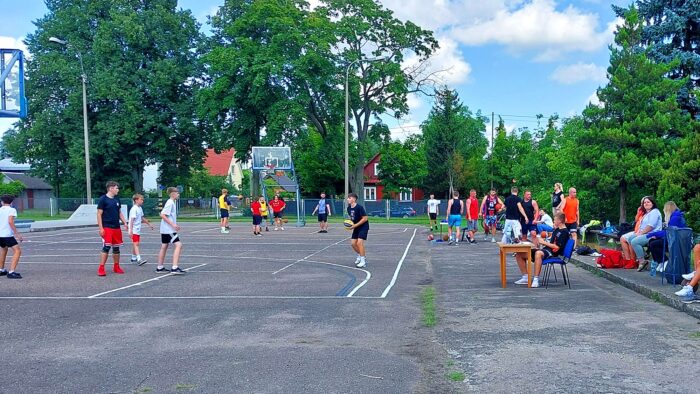 Asfaltowe boisko do gry w koszykówkę uliczną. Na nim grające dzieci, po lewej trybuny z kibicami.