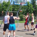 Młodzi mężczyźni w sportowych strojach, grający w koszykówkę na asfaltowym boisku.