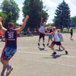 Młodzi mężczyźni w sportowych strojach, grający w koszykówkę na asfaltowym boisku.