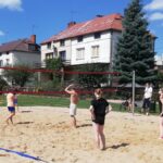 Młodzież grająca w siatkówkę plażową na boisku w parku. W tle domy jednorodzinne.