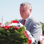 Burmistrz z poważną miną niosący wieniec przewiązany biało-czerwoną wstęgą