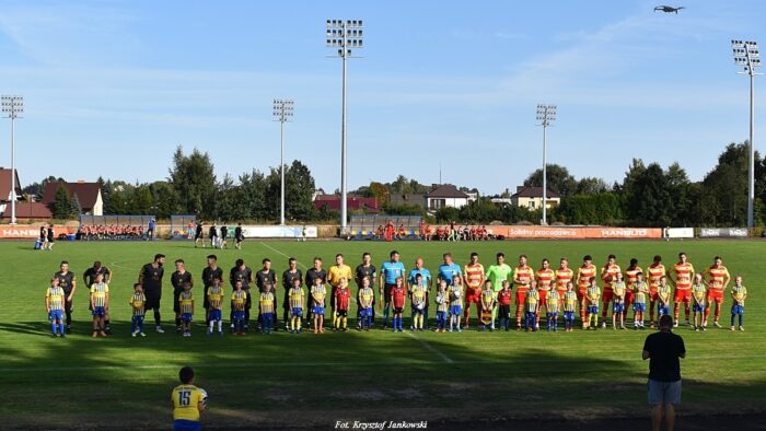 Drużyny piłkarskie Tura i Jagiellonii przed meczem. Piłkarze stoją w rzędzie, przed nimi młodzież klubowa, dzieci w strojach piłkarskich.