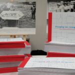 Stos książek wydanych w ramach projektu leży na tle ram z prezentowanymi zdjęciami historycznymi.