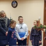 Troje dzieci kolędników stoi wraz z dwiema nauczycielkami. Obok burmistrz ze stroikiem świątecznym w dłoniach.