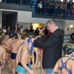 Burmistrz nakłada medale na szyje kolejno podchodzącym do niego młodym zawodnikom. W tle trybuny pływalni i kibice.