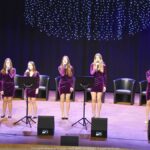 Zespół wokalny - osiem młodych kobiet - na scenie śpiewa do ustawionych mikrofonów.