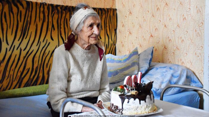 Uśmiechnięta starsza kobieta z chustką na łowie siedzi na łóżku. Przed nią stoi tort ze świeczkami tworzącymi napis "100".