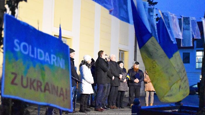 Na pierwszym planie niebiesko-żółte flagi Ukrainy oraz wywieszone zdjęcia. W tle grupa osób stojąca pod ratuszem.