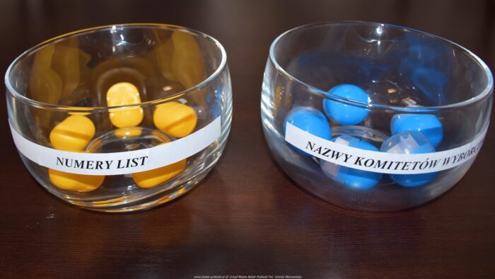 Dwa szklane pojemniki wypełnione kulkami - jeden w kolorze żółtym, drugi w niebieskim., które posłużyły losowaniu. Na lewym pojemniku napis "numery list", na prawym napis "nazwy komitetów wyborczych".