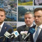 Trzech mężczyzn podczas konferencji prasowej na tle ścianki miasta Bielsk Podlaski z kolorowymi zdjęciami. Do mikrofonu przemawia jeden z mężczyzn - minister finansów.