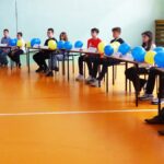Młodzi uczestnicy konkursu siedzą za stołami ustawionymi w łuk. Stoły przyozdobione są niebiesko-żółtymi balonami.