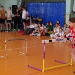 Dziewczyna biegnie wokół płotka pokonując trasę konkurencji sportowej. W tle inni uczniowie obserwujący zawody na sali gimnastycznej.
