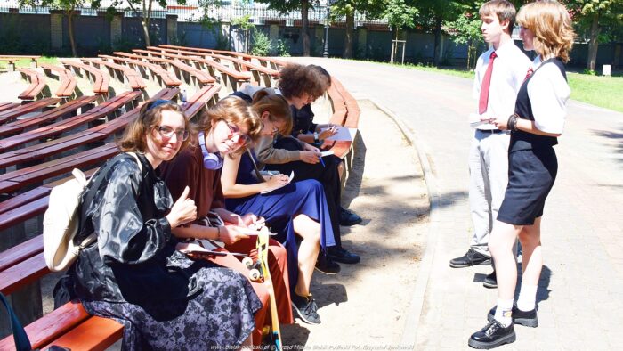 Grupa młodzieży siedzi na drewnianych ławkach amfiteatru. Wypełniają ankiety. Jedna z pań pokazuje do aparatu gest - uniesiony kciuk. Obok stoją dwie osoby z grupy inicjatywnej Młodzieżowej Rady Miasta.