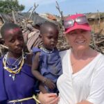 Nauczycielka z Bielska Podlaskiego trzyma dziecko z Kenii na rękach. Obok stoi kobieta z Kenii. W tle afrykańskie lepianki.