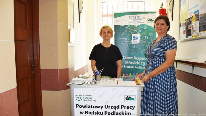 Dwie kobiety stoją przy stoisku na korytarzu urzędu. Przed nimi stolik z gadżetami promocyjnymi i napisem "Powiatowy Urząd Pracy w Bielsku Podlaskim", a za nimi roll-up reklamujący Centrum Wspierania Młodzieży.