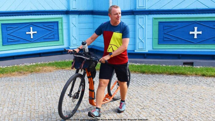 Kolorowo ubrany mężczyzna z hulajnogą na tle ściany niebieskiej cerkwi.