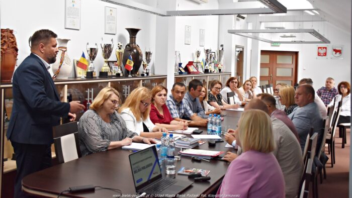 Grupa ludzi siedzących za długim stołem w trakcie spotkania i dyskusji. Po prawej burmistrz mówiący i gestykulujący do zebranych.