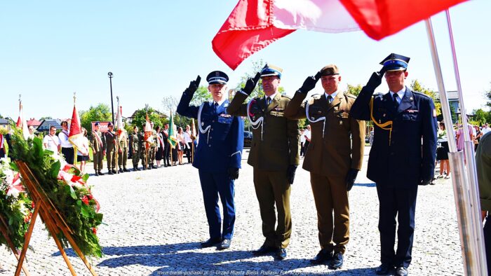 Czterech mężczyzn w mundurach oddaje hołd salutując przed pomnikiem. Nad nimi powiewają biało-czerwone flagi. W tle widać poczty sztandarowe i uczestników uroczystości.