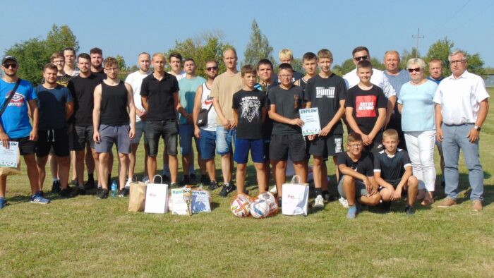 Grupowe zdjęcie uczestników i organizatorów zawodów. Młodzież oraz dorośli stoją na boisko i pozują do fotografii.