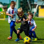 Sześcioletnie dzieci w strojach sportowych grają w piłkę na zielonej sztucznej murawie bielskiego stadionu.