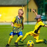 Sześcioletnie dzieci w strojach sportowych grają w piłkę na zielonej sztucznej murawie bielskiego stadionu.