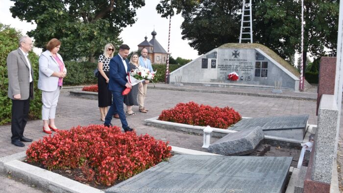 Kilkuosobowa delegacja urzędników stoi przed pomnikiem. W jego kierunku idzie burmistrz z wieńcem biało-czerwonych kwiatów. W tle inne pomniki i cmentarz.